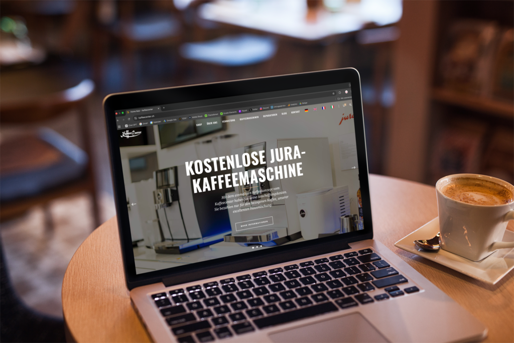 A MacBook on a café table displaying the Kaffeecenter GmbH homepage and the title ‘Free Jura coffee machine’