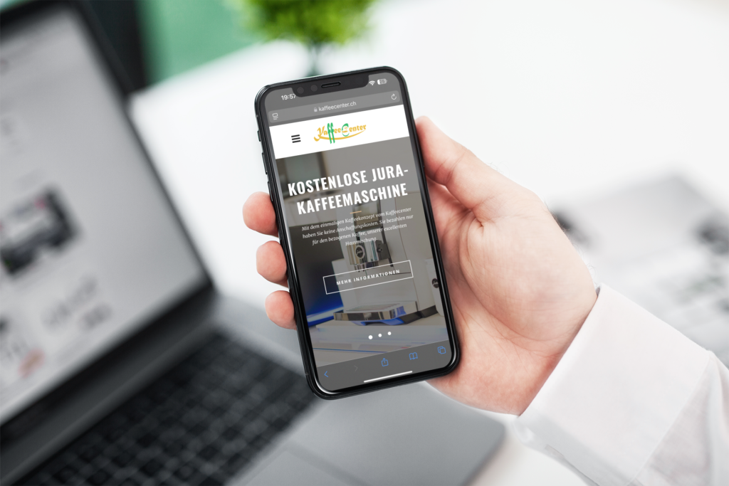 A hand is holding an iPhone displaying the Kaffeecenter GmbH mobile homepage, with a laptop behind it