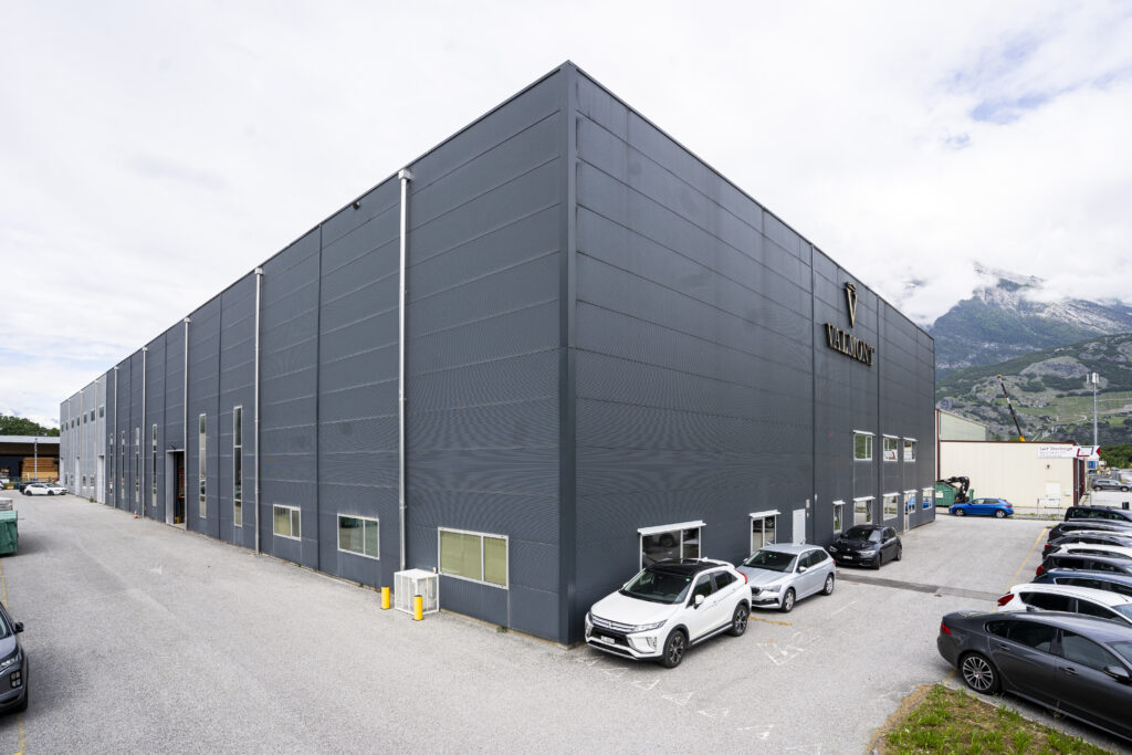The rear corner of the dark Valmont hall in Vétroz, with the logo at the right-hand end