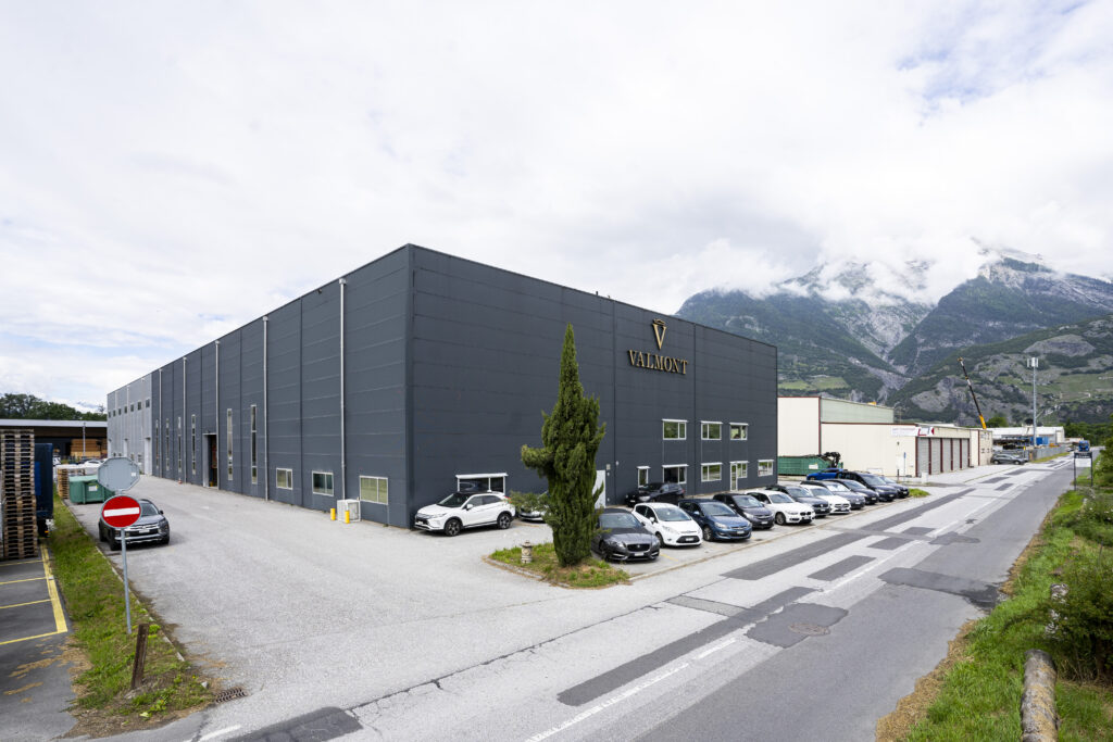 The long, dark façade of the Valmont hall in Vétroz, seen from the street corner, with the Valais mountains behind it