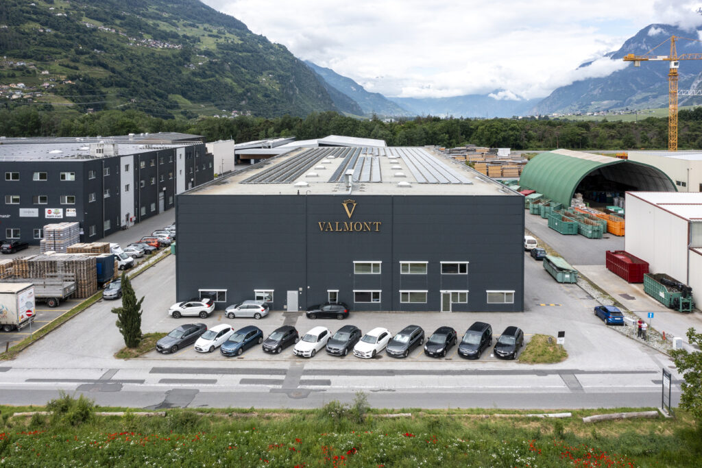 A dark industrial hall bearing the Valmont logo in Vétroz, with a row of cars in front of it and mountains behind it
