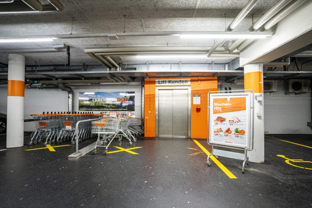 Car park level in Zuchwil, featuring the customer lift, shopping trolleys and a Migros poster