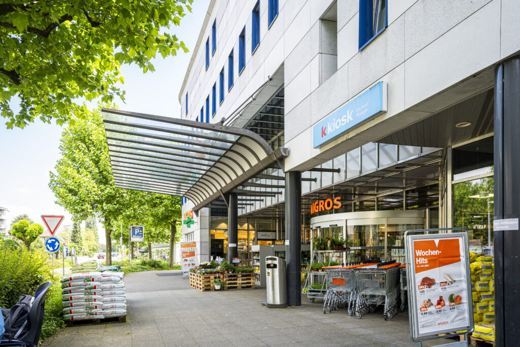 The entrance to Migros in Zuchwil under the glass canopy, with the k-kiosk and shopping trolleys next to it