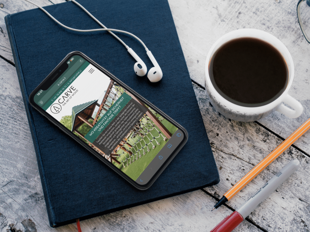 An iPhone on a notebook featuring the Carve website and a wooden play tower, with coffee and pens next to it