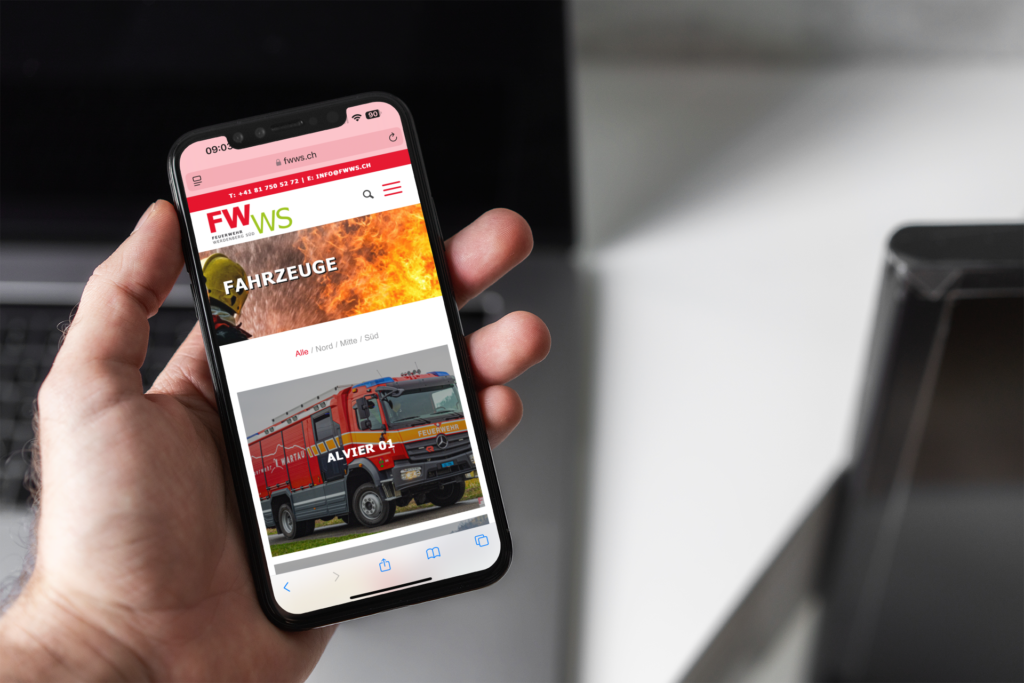 A hand holding an iPhone showing the vehicles page of the Feuerwehr Werdenberg Süd website and the vehicle Alvier 01