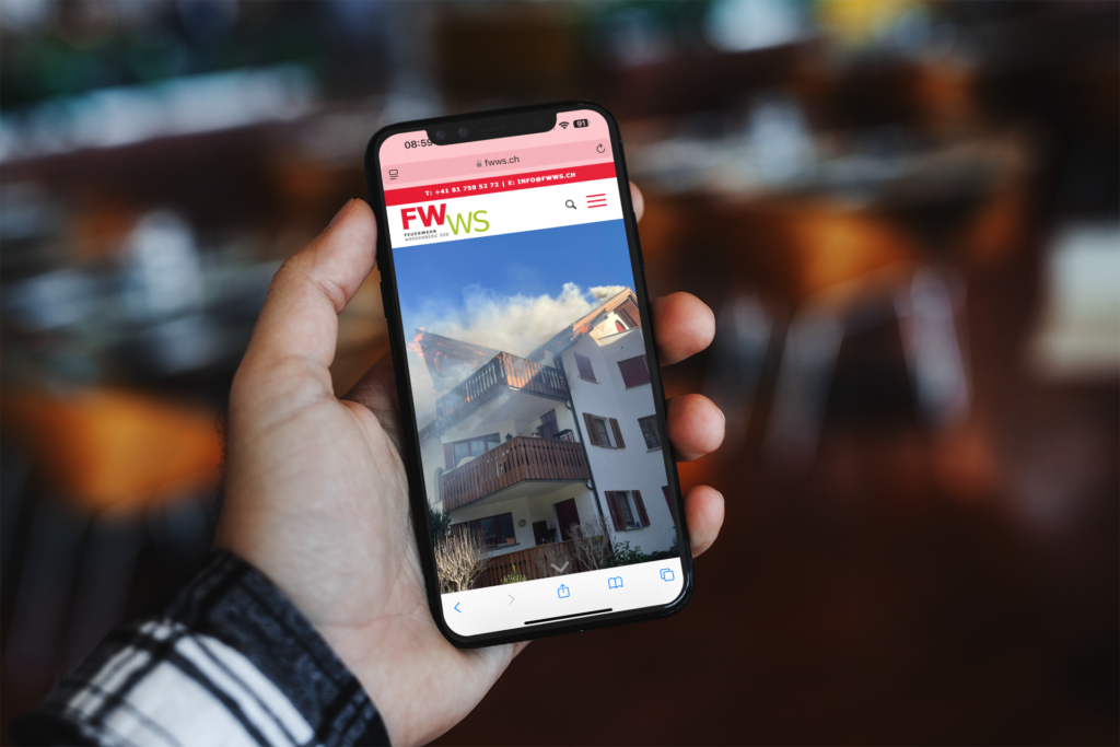 A hand is holding an iPhone displaying the Feuerwehr Werdenberg Süd website and a burning attic