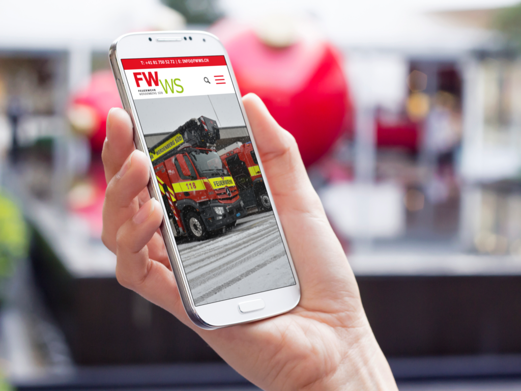 A hand is holding a smartphone displaying the mobile website of the Feuerwehr Werdenberg Süd and a red fire engine