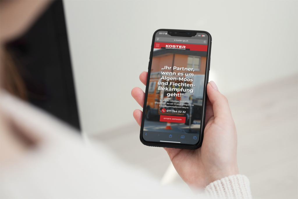 A hand is holding an iPhone displaying the mobile website of Koster Gebäudesanierung and the telephone number